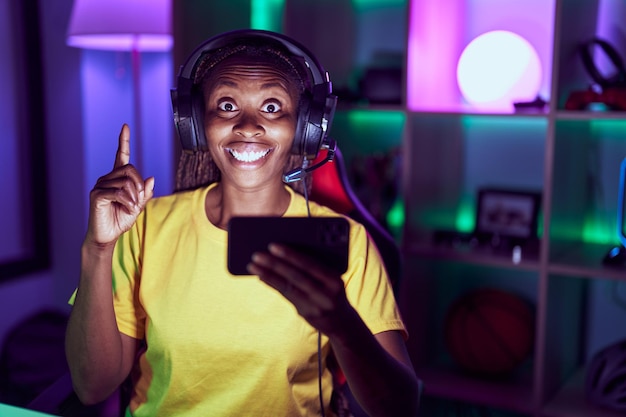 African american woman playing video games with smartphone surprised with an idea or question pointing finger with happy face number one