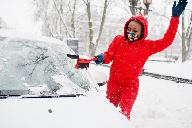 African american woman in red hoodie and face mask clean car from snow in winter day.