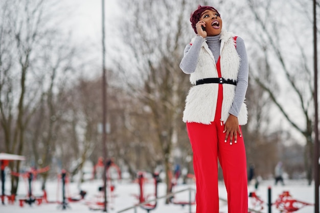 African american woman in red pants and white fur coat jacket posed at winter day against snowy background speaking on phone