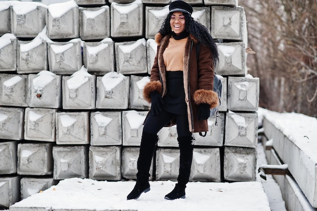 African american woman in sheepskin coat and cap posed at winter day against snowy stone background