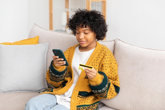 African american woman shopping online holding smartphone paying with gold credit card girl sitting