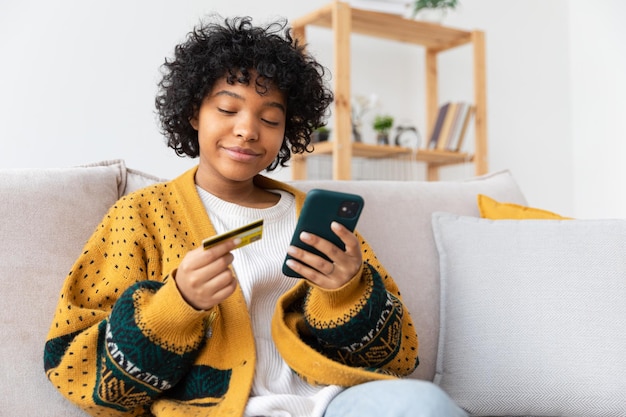 African american woman shopping online holding smartphone paying with gold credit card girl sitting