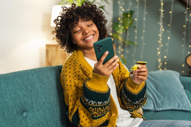 African american woman shopping online holding smartphone paying with gold credit card girl sitting