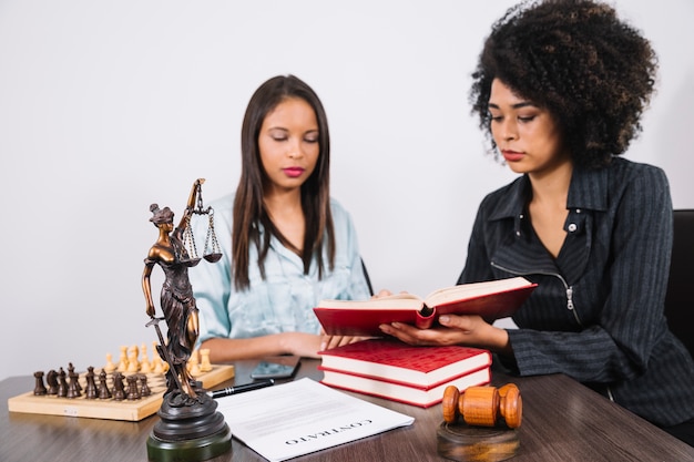 African American woman showing book to lady at table with document, smartphone and chess