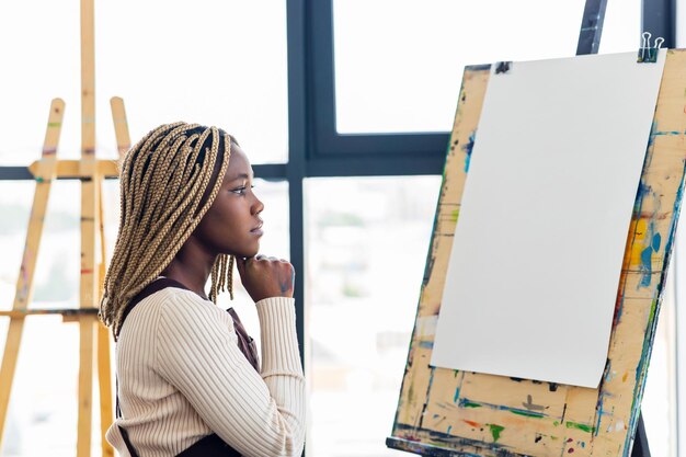 African american woman sitting next to blank white canvas on easel in workspace