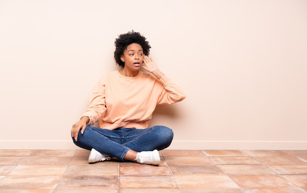 African american woman sitting on the floor listening to something by putting hand on the ear