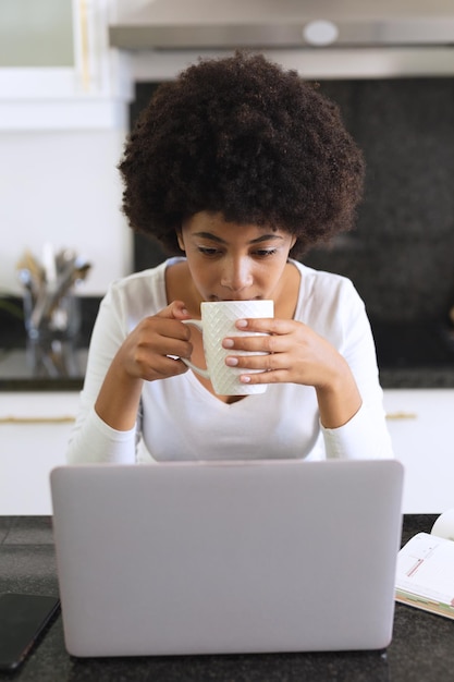 キッチンに座ってコーヒーを飲み、ラップトップを使用しているアフリカ系アメリカ人の女性