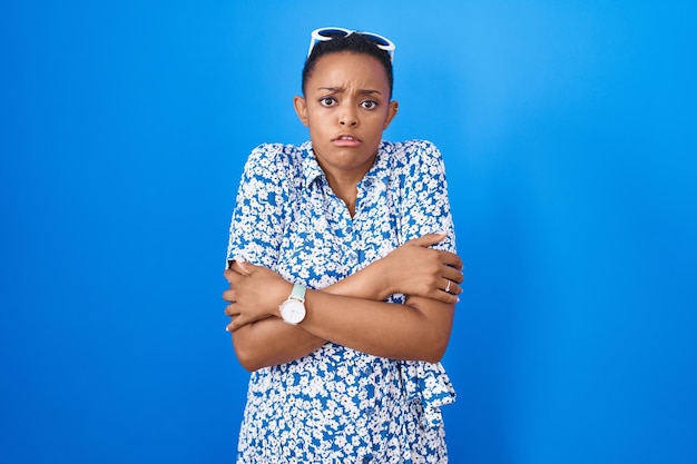 African american woman standing over blue background shaking and freezing for winter cold with sad and shock expression on face