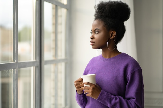 African American woman thinking, looking through the window, holding mug, drinking coffee