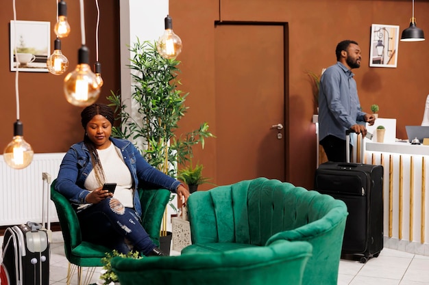 アフリカ系アメリカ人の女性旅行者がホテルのロビーに座り、ホテルのチェックインを待ちながらスマートフォンで友人とオンラインチャットをしている。荷物を持った観光客が受付エリアで待つ