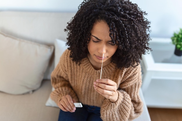 집에서 코로나바이러스 PCR 테스트를 하는 동안 면봉을 사용하는 아프리카계 미국인 여성. 코로나바이러스 신속 진단 테스트를 사용하는 여성. 집에서 COVID-19에 비강 면봉을 사용하는 젊은 여성.