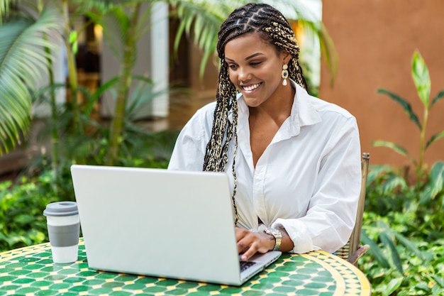 African American woman using laptop in a cafe store outside. Woman with braids sitting using laptop and drinking coffee.