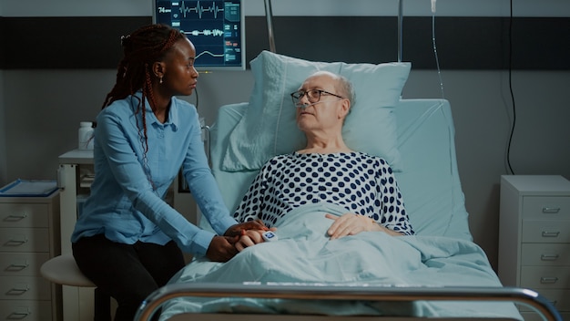 African american woman visits sick patient in hospital ward bed at medical facility old man with oxi...