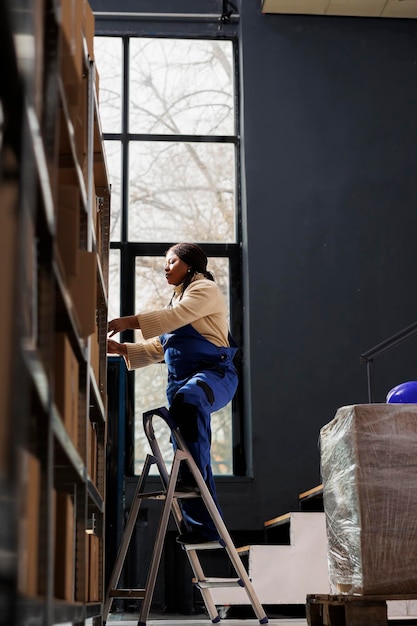 사진 아프리카계 미국인 여성 창고 노동자가 사다리 위에 서서 높은 선반에서 마분지 상자를 꺼냅니다. 물류 창고 보관실에서 고객 소포에 도달하는 우체국 직원
