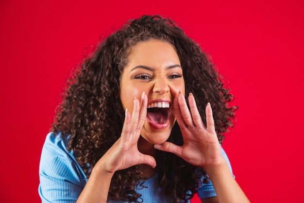 African american woman wearing a blue t-shirt over isolated red background shouting and screaming loud to side with hand on mouth. Communication concept.