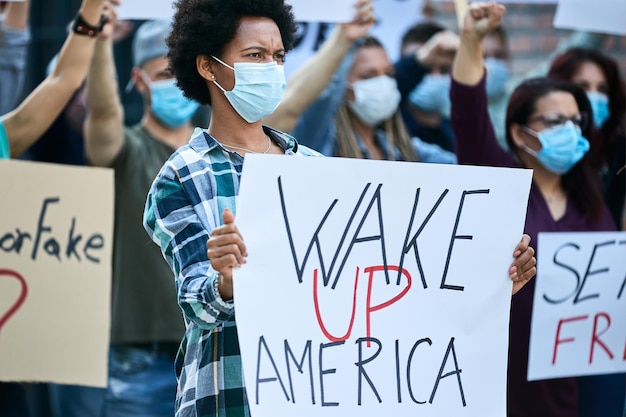 African American woman wearing protective face mask and carrying banner while protesting with crowd of people on city streets