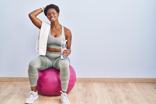 African american woman wearing sportswear sitting on pilates ball smiling confident touching hair with hand up gesture, posing attractive and fashionable