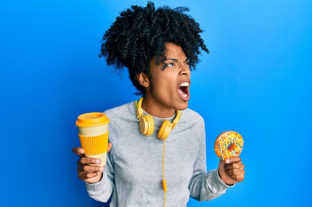 African american woman with afro hair eating doughnut and drinking coffee angry and mad screaming frustrated and furious, shouting with anger. rage and aggressive concept.