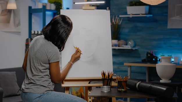 African american woman with artist occupation drawing vase on white canvas using pencil while sitting in creativity studio. Black young artist working on art masterpiece as artistic hobby