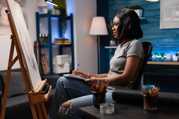 African american woman with artistic abilities appreciating\
completed pencil drawing in modern art studio. female professional\
artist revising artwork to give final details sitting in work\
room.