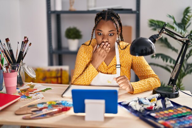 African american woman with braids sitting at art studio painting looking at tablet covering mouth with hand shocked and afraid for mistake surprised expression