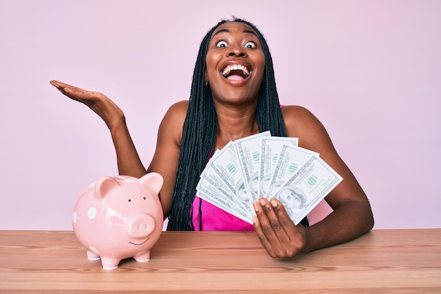 African american woman with braids sitting on the table with piggy bank and dollars celebrating victory with happy smile and winner expression with raised hands