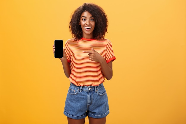 African American woman with curly hair pointing at cellphone over orange wall