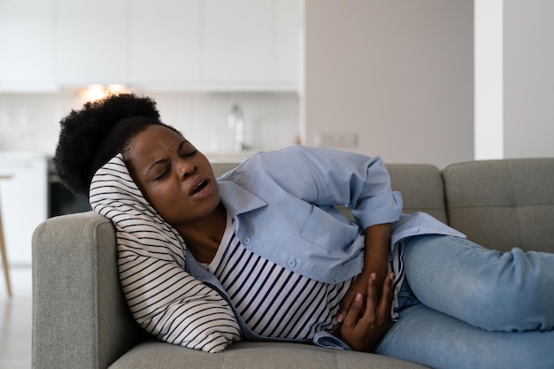 African American woman with health problems suffering from cramps and stomach pain lies on sofa