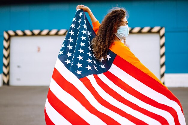 Donna afroamericana con maschera facciale medica che posa con la bandiera americana concetto di prevenzione