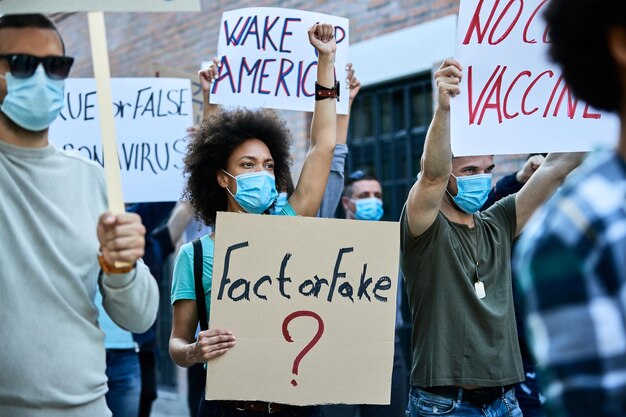 African American woman with raised fist wearing protective face mask while carrying banner with 'fact or fake' inscription during public demonstrations