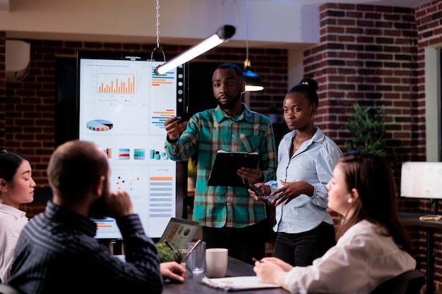 African american work colleagues brainstorming marketing strategy with sales team working overtime in late night meeting. Two coworkers discussing business ideas with board members in startup office.