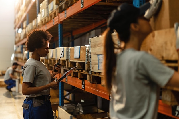 Афроамериканский рабочий читает график доставки, разговаривая по рации в промышленном складском помещении
