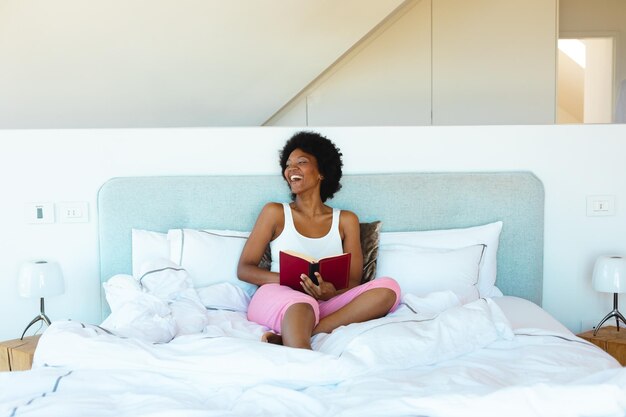 Африканская молодая афроамериканка смеется, сидя дома с книгой на кровати, копирует пространство. Неизменное, хобби, образ жизни и концепция дома.