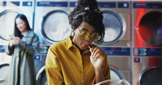 African American young beautiful girl in yellow glasses sitting in laundry service room. Woman with lollypop reading magazine while waiting for clothes to be washed. Female clients .