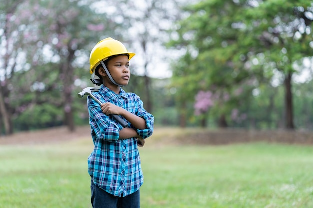 写真 アフリカ系アメリカ人の少年がスチールハンマーを押しながら屋外に立っている間安全メガネと黄色の安全ヘルメットを着用します。