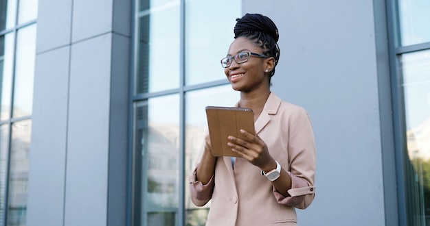 African American young businesswoman in glasses tapping and scrolling on tablet device. Woman texting message on computer outdoors. Confident female using gadget and chatting.