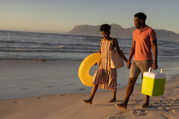 Photo african american young couple with cooler and inflatable ring holding hands while walking on beach