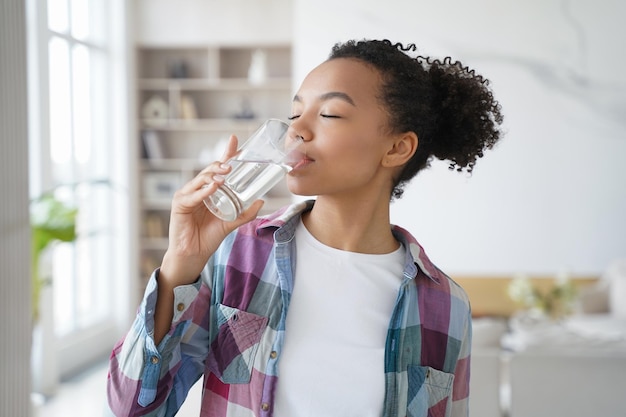 アフリカ系アメリカ人の若い女の子は家でガラスから純粋なろ過水を飲むことを楽しんでいます健康的なライフスタイル