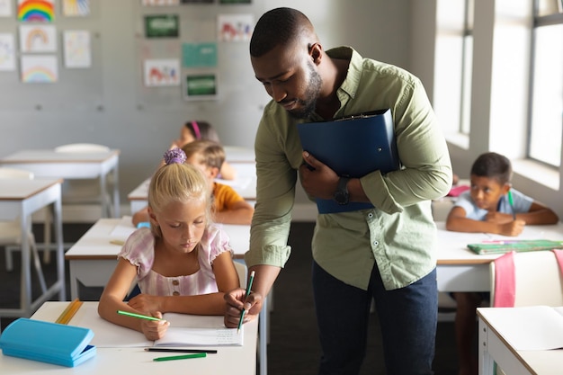 아프리카계 미국인 젊은 남성 교사가 교실에서 책상에 앉아있는 백인 초등학교 소녀를 돕고 있습니다. 변경되지 않은 교육, 학습, 어린 시절, 직업, 교육 및 학교 개념.