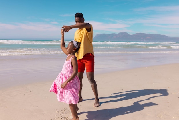 晴れた日に海と空を背景にビーチで幸せな娘と踊るアフリカ系アメリカ人の若者