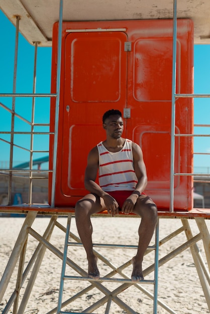 African american young man looking away while sitting on lifeguard hut at beach on sunny day