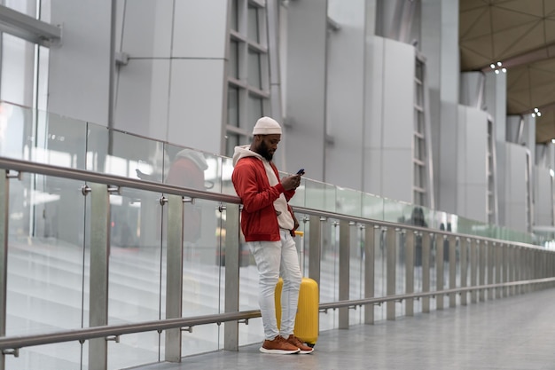 空港ターミナルで荷物を持って旅行するスマートフォンを使用してアフリカ系アメリカ人の若い男