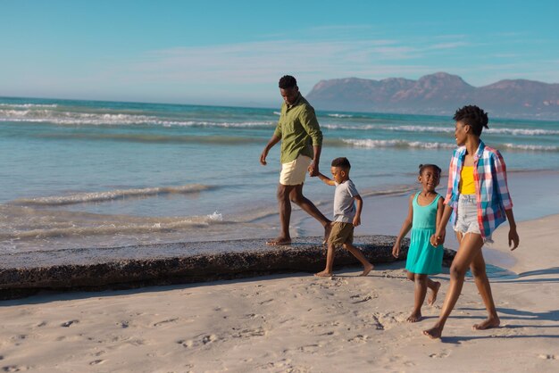 African american young parents holding daughter and son's hands while walking at beach against sky