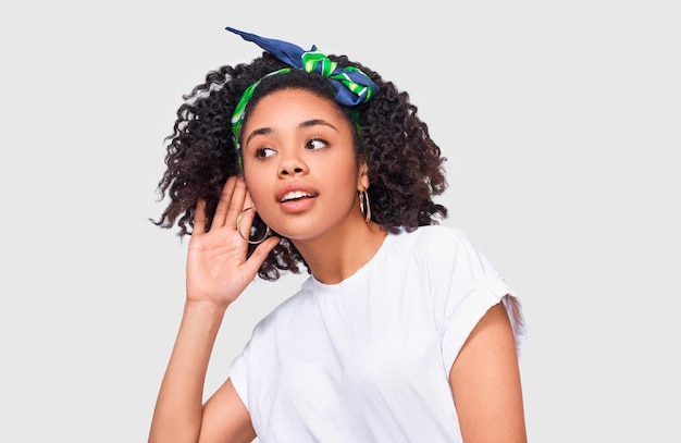 Photo african american young woman paying attention and placing hand on ear asking someone to speak louder or whisper isolated on white wall pretty afro girl which overhears secret conversation