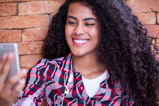 Studente di giovane donna afroamericana che fa selfie foto sulla fotocamera anteriore dello smartphone per la condivisione in reti seduto sul pavimento