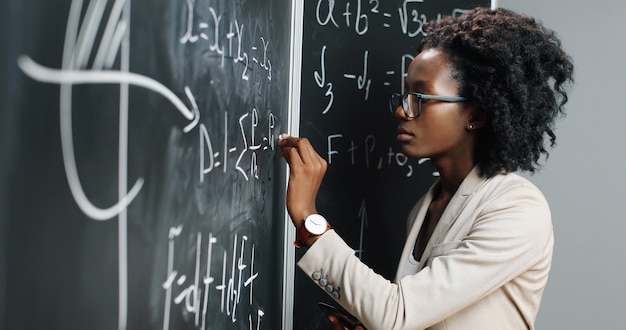 African American young woman teacher at school writing formulas and mathematics laws on blackboard. School concept. Female math lecturer in glasses explaining physics laws. Education.