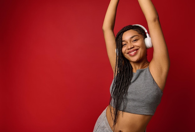 African American young woman with wireless headphones in gray sportswear enjoys listening music, dances with raised arms, smiles toothy smile looking at camera against red background with copy space