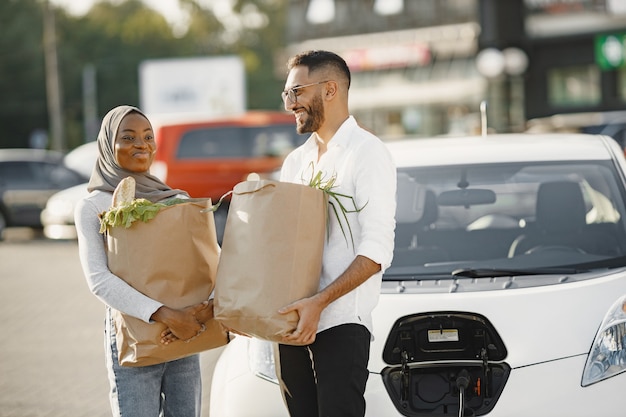 La coppia araba africana sta con generi alimentari vicino all'auto elettrica. ricarica dell'auto elettrica alla stazione di servizio elettrica