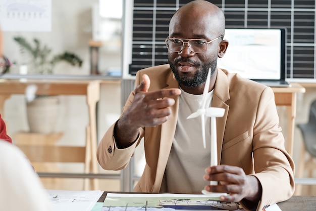 African architect pointing at model of windmill in his hand and presenting it to his colleagues