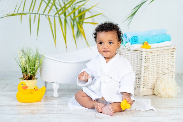 Un bambino africano in camice bianco dopo il bagno e il bagno gioca con un'anatra a casa, il concetto di cura e igiene dei bambini piccoli.